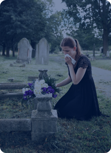 Woman in black dress kneeling and crying at a grave, holding a tissue beside a bouquet of flowers in a cemetery.